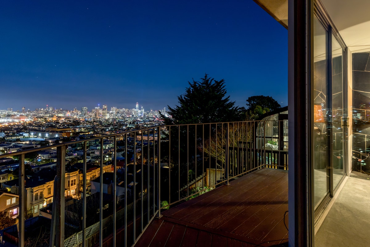 A spacious balcony offers a panoramic nighttime view of the city skyline, with twinkling lights reflecting off the buildings. The railing provides safety while an adjacent glass door allows natural light to brighten the interior space.