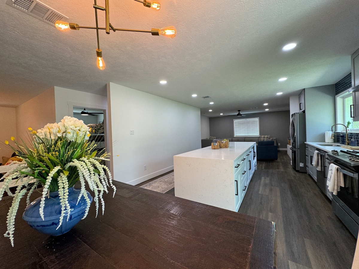 A modern kitchen layout is showcased, featuring a central island with a white countertop and ample storage beneath. Bright lights illuminate the space, complemented by a sleek dining area in the background. A decorative vase with flowers adds a touch of greenery to the scene.