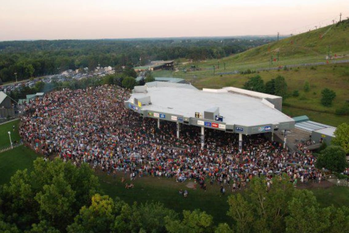 A large outdoor venue is seen filling with a diverse crowd, gathered on a lush green lawn. The venue's architecture features a wide overhang, with bright signage visible under a clear sky, while hills are visible in the background.