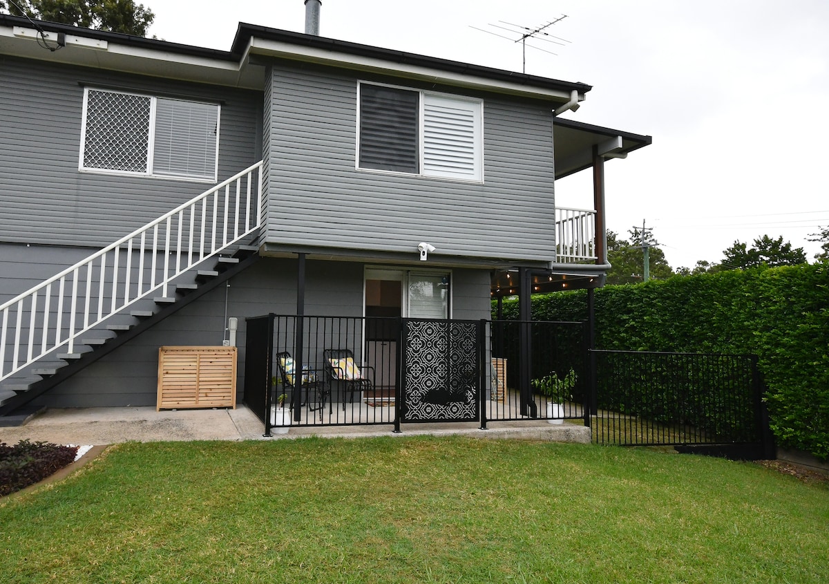 The exterior view showcases a modern two-story structure, featuring a private entrance with an accessible fenced area. A well-maintained yard with green grass is visible, complemented by decorative fencing and a set of outdoor chairs. The upper level includes a staircase leading up.