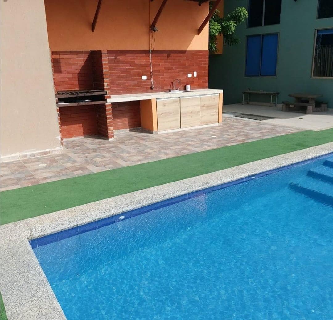 A modern outdoor area features a clear swimming pool surrounded by light-colored stone. An outdoor kitchen with a brick grill and wooden counter is visible, alongside a green lawn. A wooden picnic table is positioned nearby, enhancing the communal space.