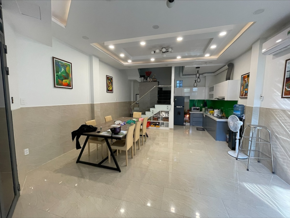 An open-layout dining area features a rectangular wooden table surrounded by beige chairs. Brightly colored artwork adorns the walls, and a staircase leads to an upper level. A modern kitchen with green accents is visible in the background, complemented by ample lighting throughout the space.