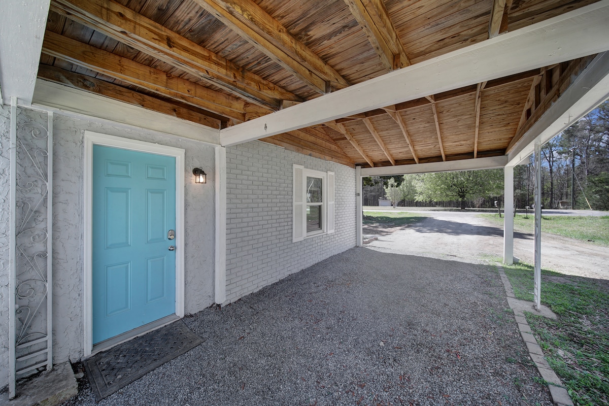 An inviting entrance features a light blue front door and is sheltered under a wooden carport. A gravel pathway leads to the door, while windows allow natural light to enter the space, creating a welcoming atmosphere.