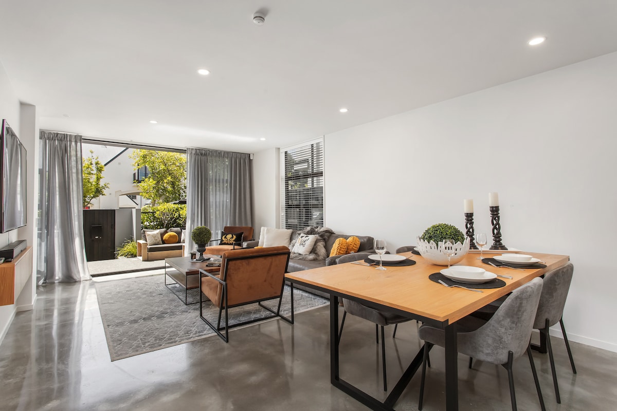 An open plan living and dining area features a wooden table set for dining, complemented by comfortable seating on a plush couch. Large sliding doors invite soft light and offer a view of a sheltered courtyard.