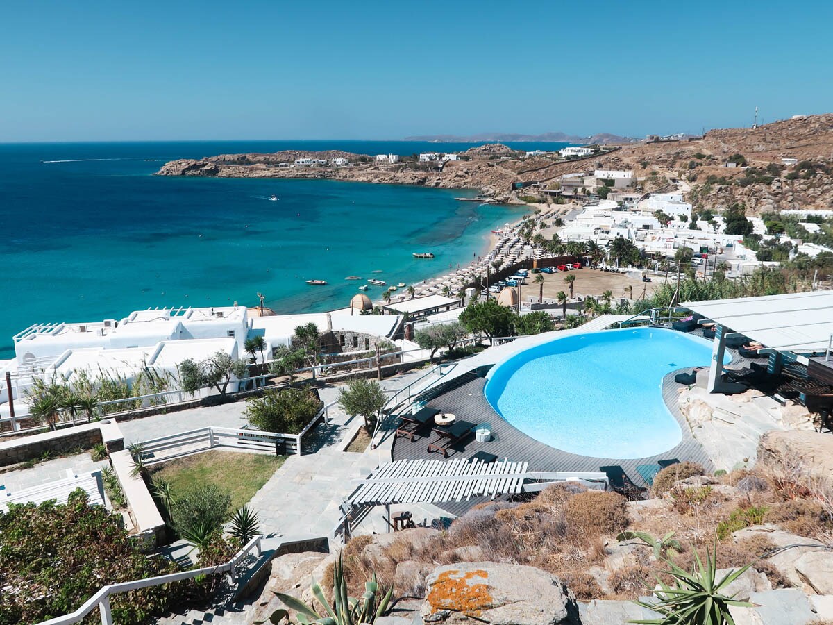 A panoramic view captures the coastline and clear blue waters, with a large infinity pool set against a backdrop of white buildings. The landscape includes sandy beaches and rugged hills, offering a sense of tranquility and connection to nature.