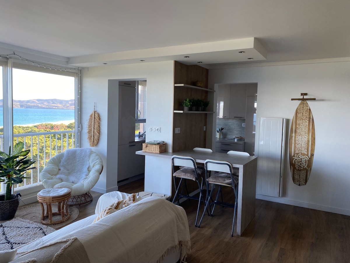 A bright living space features a cozy seating area with a large armchair and a soft throw. A small kitchen island with two stools offers a casual dining option. Large windows provide expansive views of the ocean and natural landscape outside.