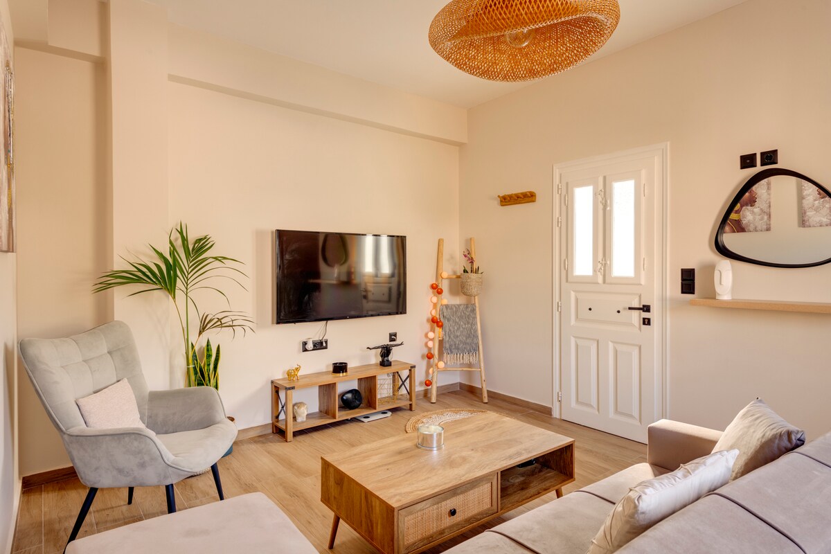 A light-filled living room features a comfortable sofa and a cozy armchair. A wooden coffee table is positioned centrally, while a wall-mounted TV is visible. Natural elements are introduced through plants, and the warm lighting enhances the room's contemporary design. The entrance is inviting with modern decor.