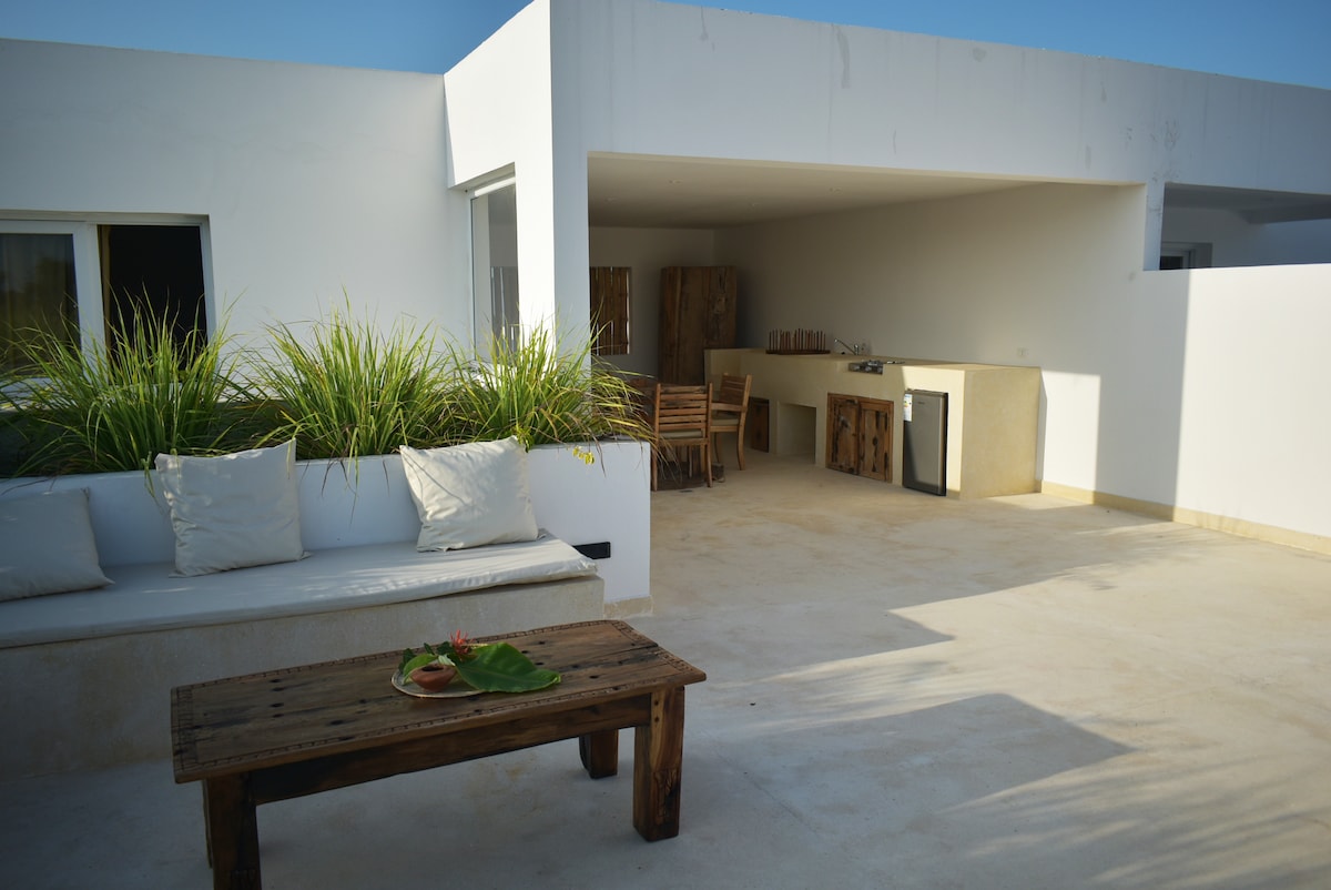 An outdoor living area features a wooden table with a small decorative arrangement and a cozy seating section surrounded by greenery. An outdoor kitchen area is visible, equipped with a fridge and countertop, all set against a bright, minimalist backdrop.