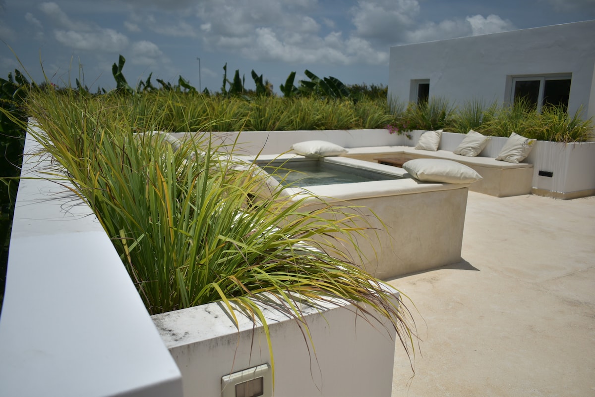A spacious terrace is surrounded by low white walls adorned with lush green grass. A small jacuzzi is positioned at one end, complemented by several large cushions arranged nearby for relaxation. The area is bathed in natural light, with a clear view of the sky.