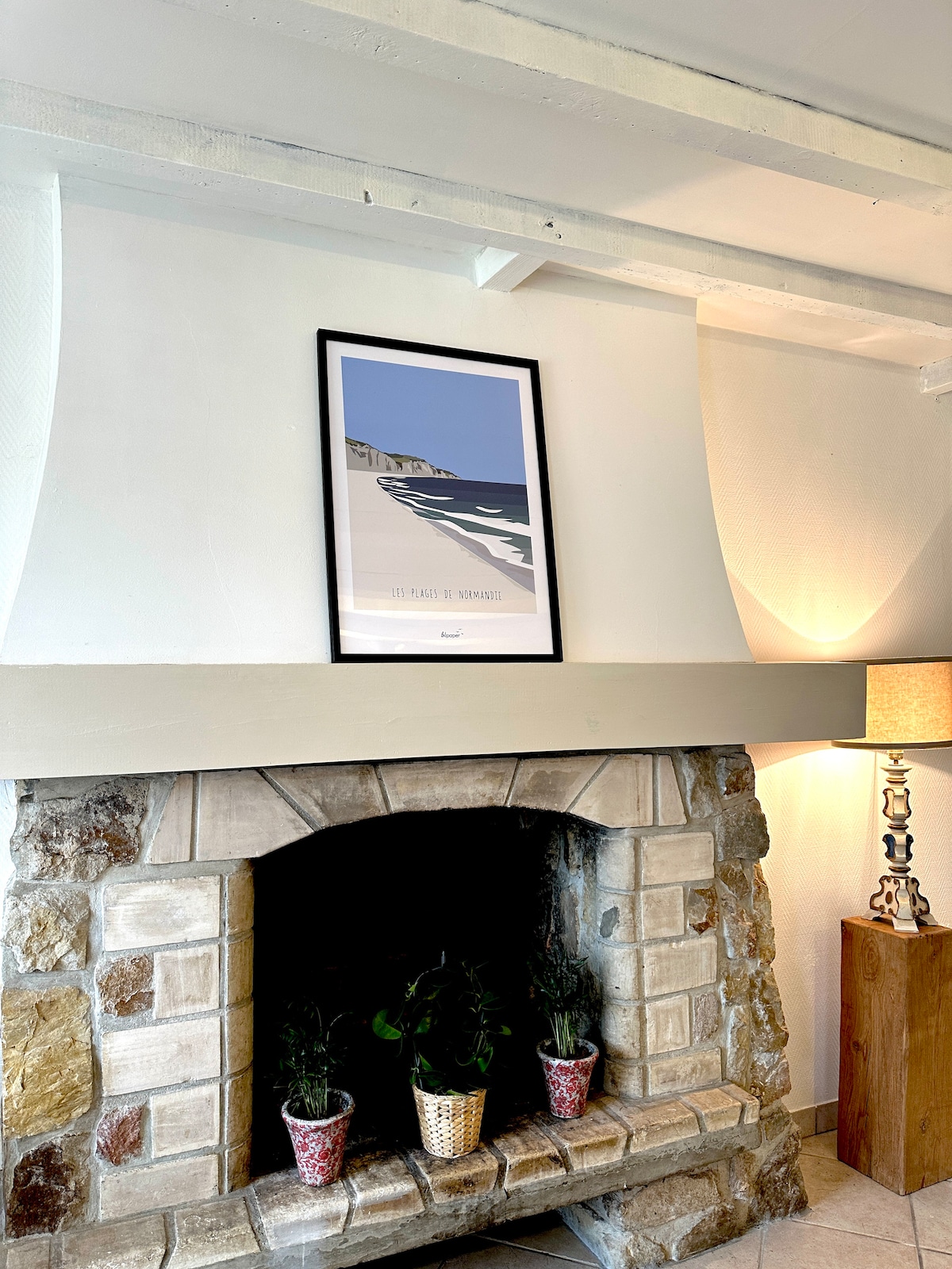 The living area features a charming stone fireplace decorated with potted plants on either side. Above the fireplace, a framed artwork depicting a beach scene is prominently displayed, while a soft lamp provides gentle illumination.