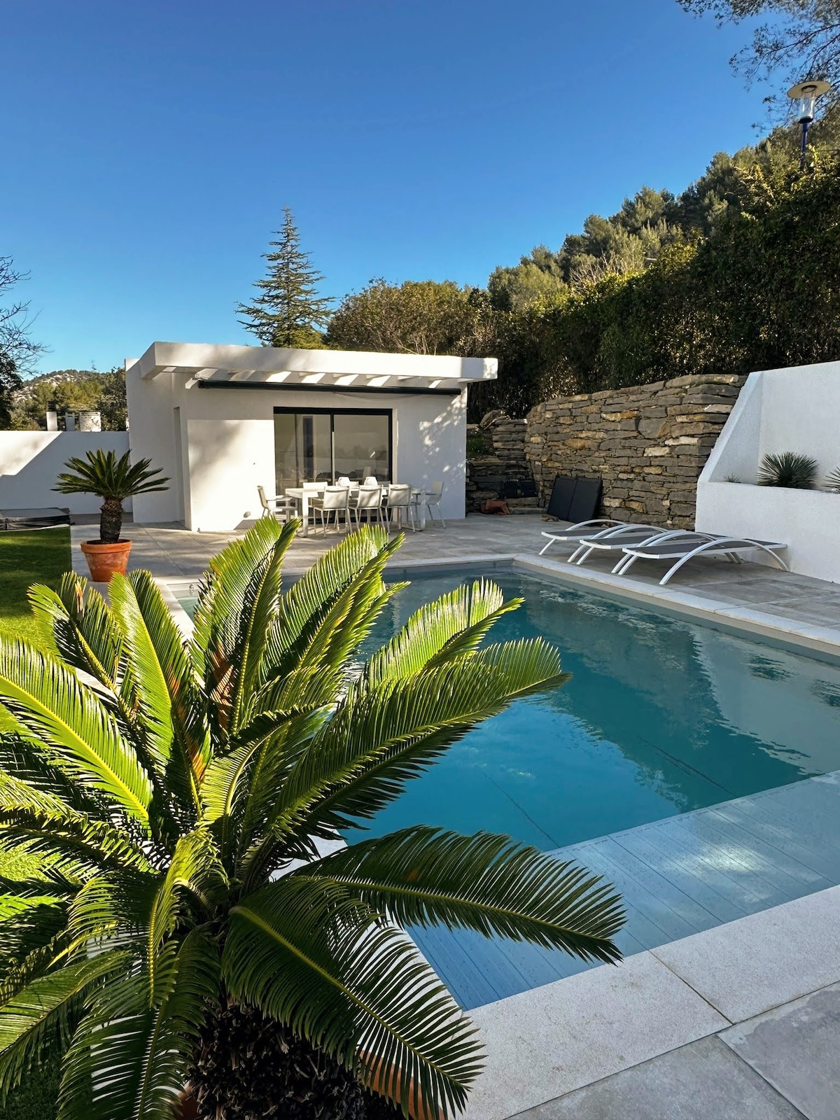 A private pool house is nestled among greenery, featuring a refreshing swimming pool bordered by sleek tiles. Sun loungers are positioned alongside, while a dining set can be seen under a shaded area. Surrounding stone walls offer a sense of seclusion.