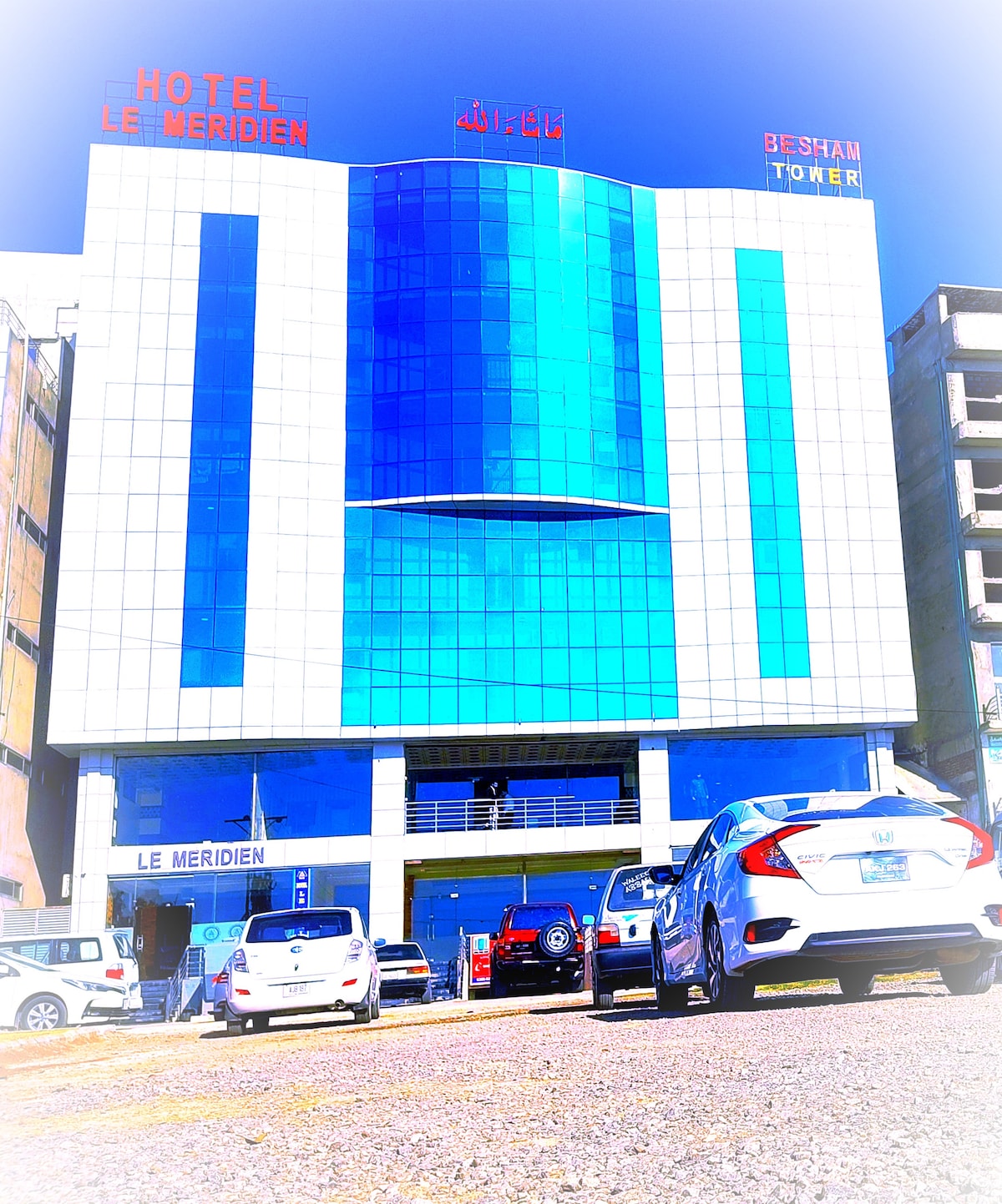 The exterior of Hotel Le Meridien is depicted, featuring a modern faรงade with large blue glass windows framed by white tiles. The hotel name is prominently displayed at the top, and parked vehicles can be seen in front, highlighting the accessible entrance.