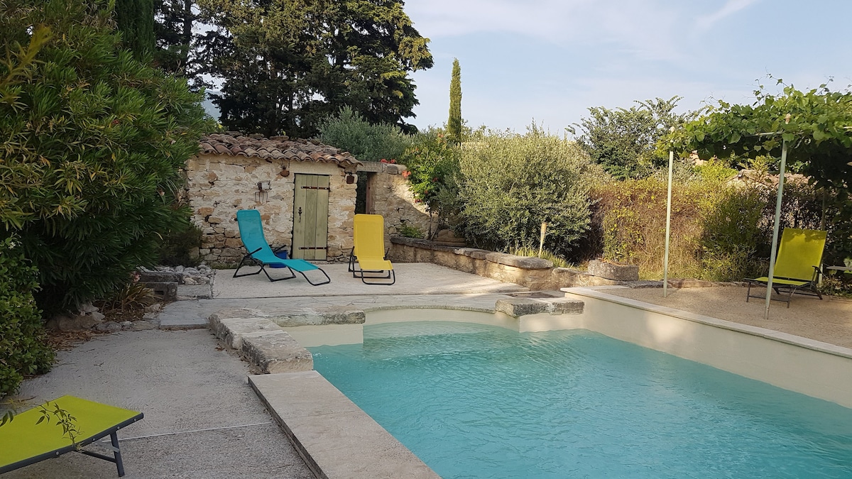 A refreshing swimming pool is surrounded by a stone terrace, featuring two lounge chairs in vibrant yellow and blue. Lush greenery and a small stone building add to the serene setting, providing a peaceful outdoor retreat.