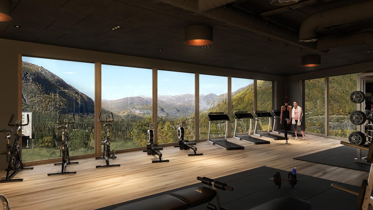 A modern gym space is equipped with various exercise machines, including stationary bikes and treadmills, arranged along large windows. Scenic mountain views are visible through the glass, enhancing the workout environment with natural light.
