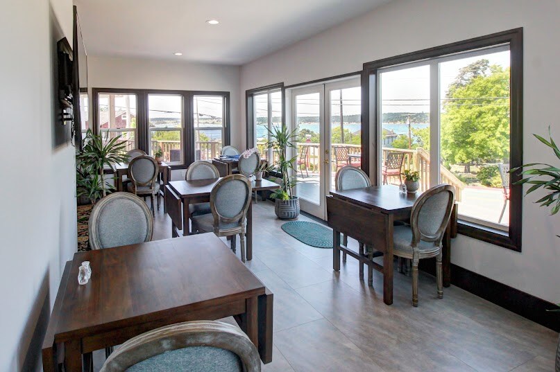 A bright dining area features multiple wooden tables surrounded by rounded chairs. Large windows allow natural light to fill the space, offering views of the outdoors. Potted plants add a touch of greenery, creating a calm atmosphere conducive to enjoying meals.