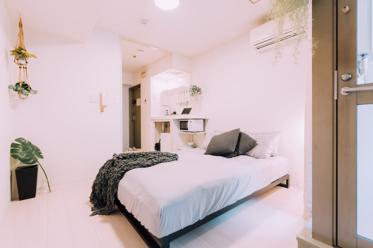 A compact studio room features a spacious bed with gray linens, positioned beside a small kitchen area that includes a microwave. A potted plant adds a touch of greenery, while light-colored walls and flooring contribute to a bright ambiance. Air conditioning is visible for comfort.