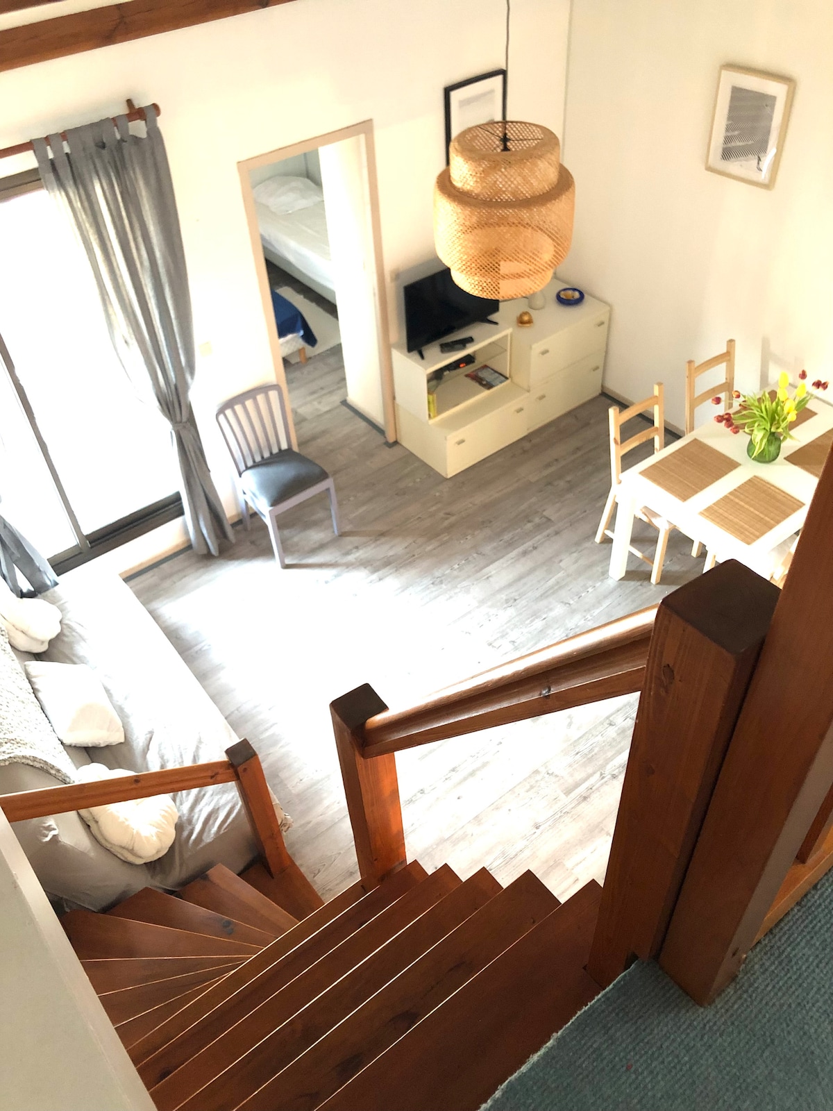 A view from the upper level shows a well-lit living space with a cozy staircase leading to the lower area. Sunlight filters through large windows, illuminating a dining table with chairs and a small television. The serene color palette is complemented by decorative elements.