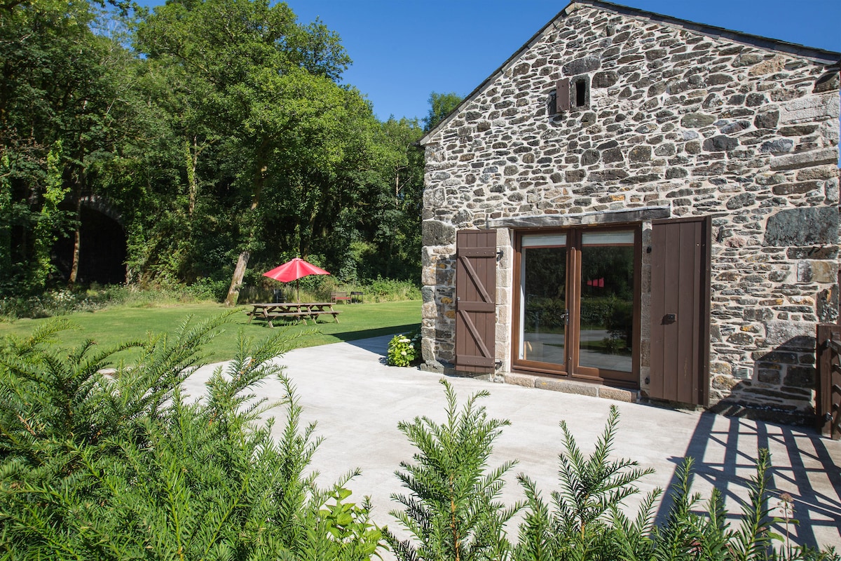A stone cottage features large windows with wooden shutters, allowing ample light. A spacious patio area is visible, bordered by greenery. A shaded picnic table is situated on the lawn, accompanied by a vibrant red umbrella, surrounded by lush trees providing a natural backdrop.