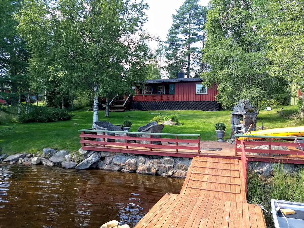 A wooden dock extends over calm waters, leading to a red cabin surrounded by lush green grass and trees. Comfortable seating arrangements are positioned nearby, creating a serene outdoor space perfect for relaxation.
