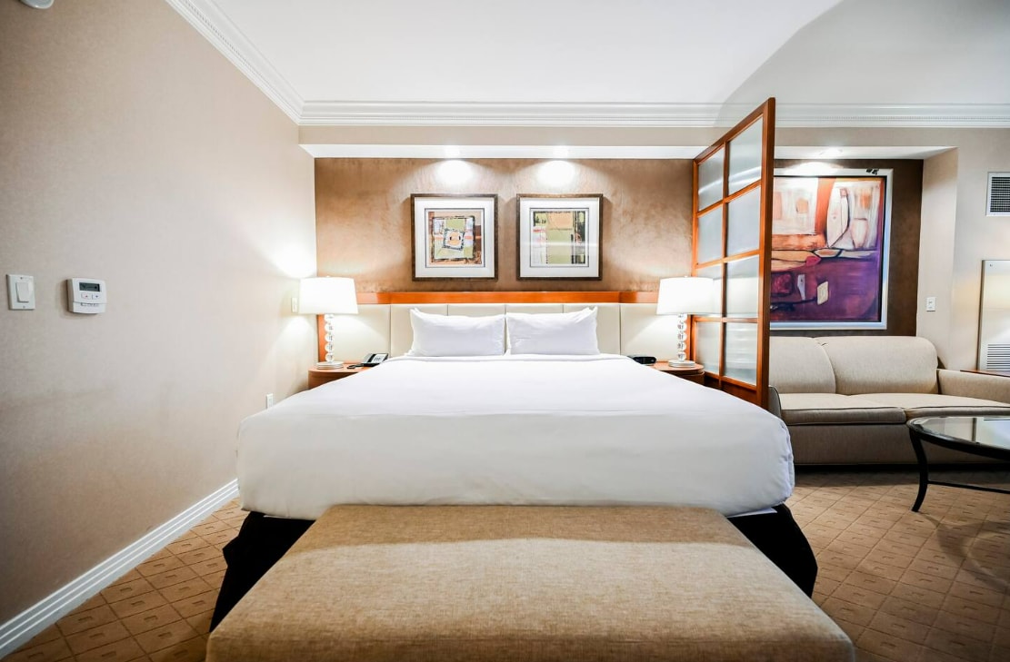 The studio features a spacious bed with a soft white duvet, framed by decorative artwork on the walls. A cozy sofa is positioned beside a glass coffee table, while light from the overhead fixtures enriches the neutral tones of the room.