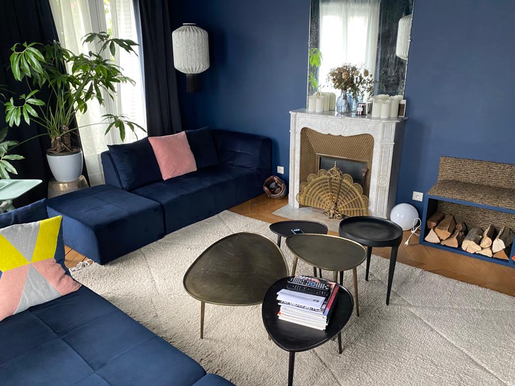 The living area features a contemporary design with a dark blue accent wall. A large sectional sofa in deep blue sits beside a stylish fireplace. Three uniquely shaped coffee tables are arranged over a soft area rug, and a potted plant adds a touch of greenery.