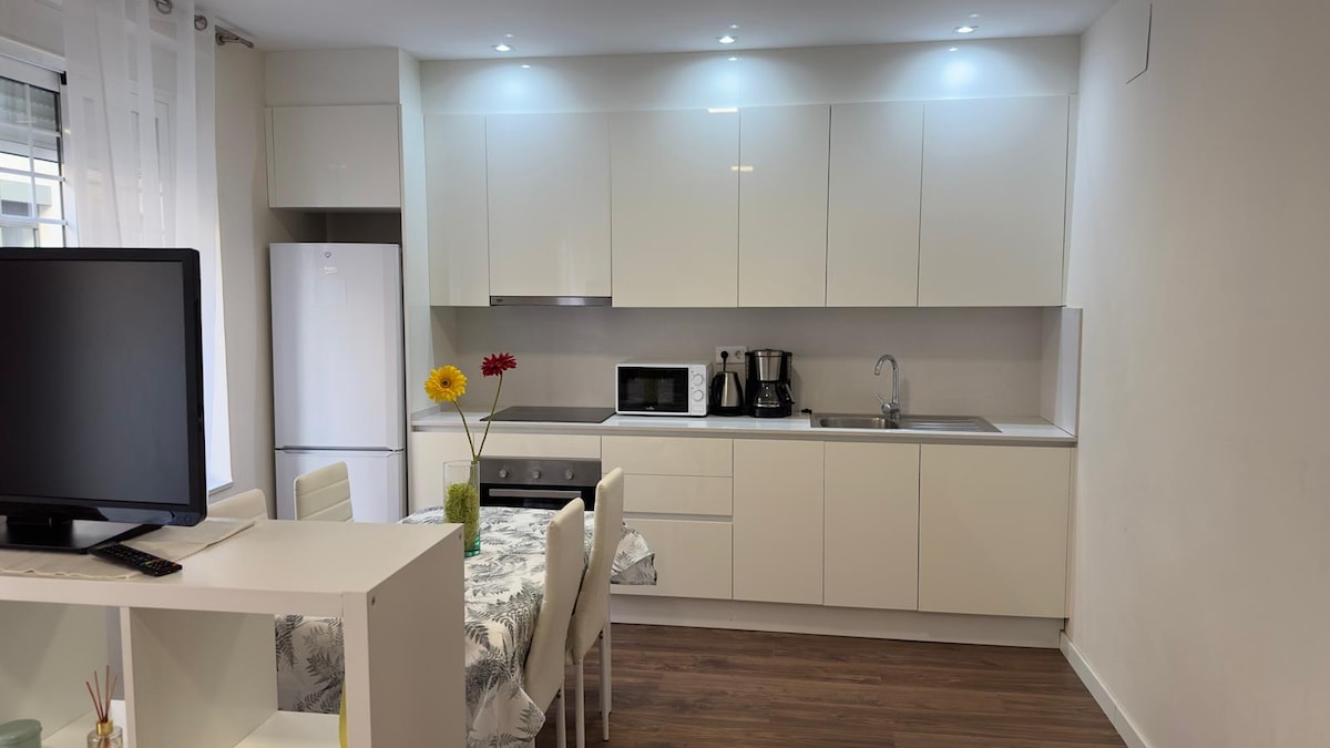 The modern kitchen features sleek white cabinetry and countertops, with a stainless-steel sink and built-in appliances including a microwave and coffee maker. A small dining table with white chairs is shown, complemented by a vase of colorful flowers. Natural light is provided by a window, enhancing the inviting space.