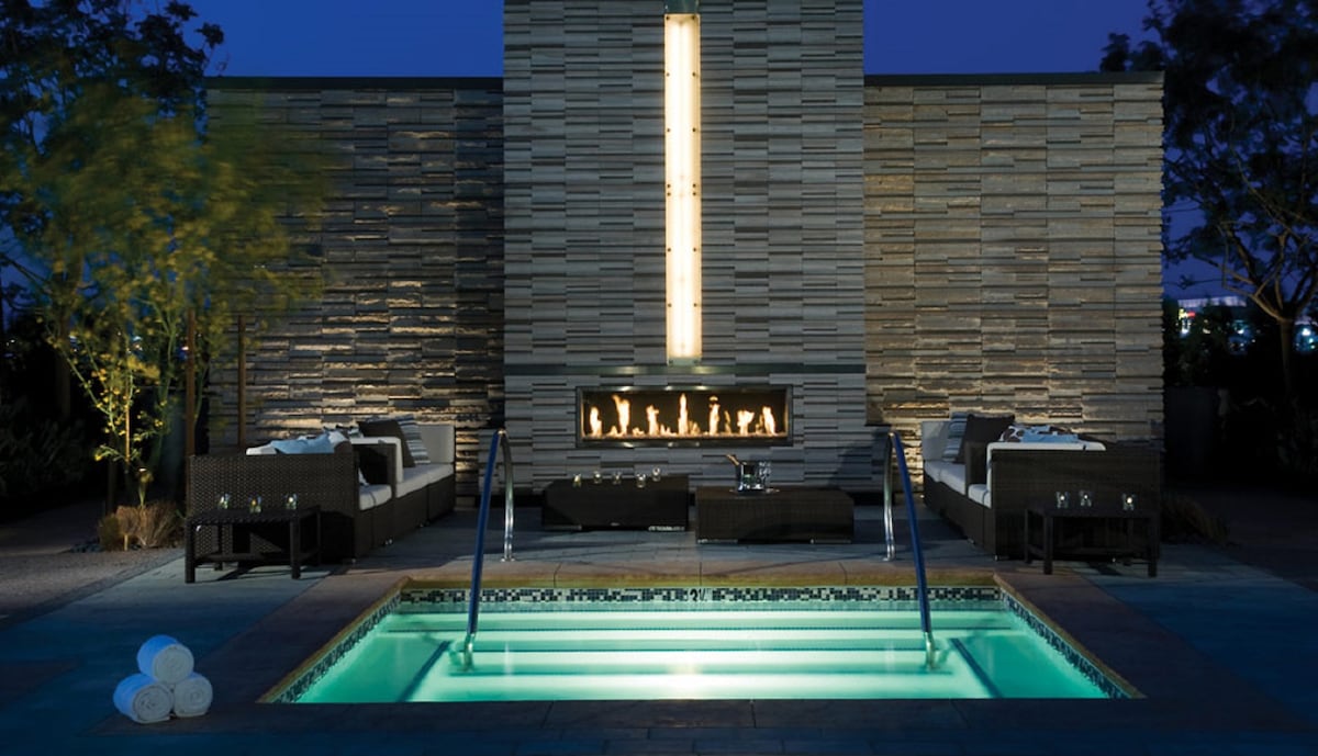 A serene outdoor space is highlighted by a warm, modern fireplace against a textured stone wall. A small, inviting pool reflects soft lighting, complemented by stylish lounge seating nearby. Light-colored towels are neatly stacked, enhancing the area’s relaxing ambiance.