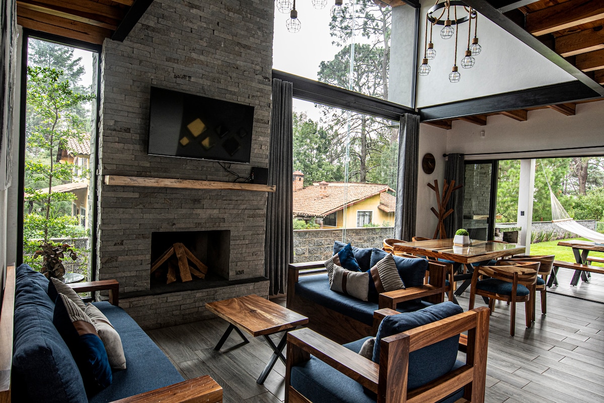 A welcoming living area features a large stone fireplace and a wall-mounted TV. Comfortable seating is arranged around a wooden table, with large windows offering views of the garden and surrounding nature. Natural light fills the space, enhancing the cozy atmosphere.