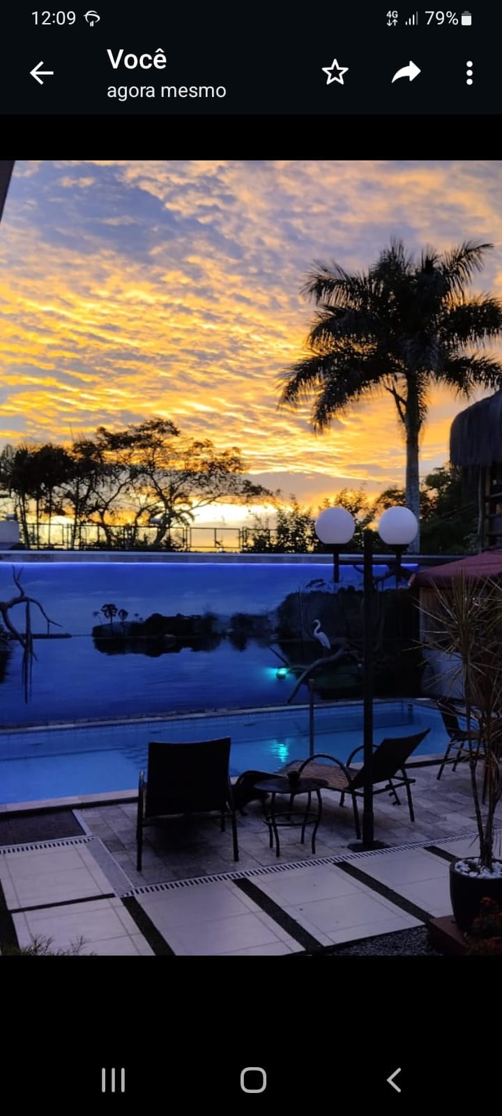 A stunning view of the swimming pool is showcased, with a backdrop of dramatic sunset colors reflected in the water. Palm trees frame the scene, and lounge chairs are arranged casually around the pool area, illuminated by soft lighting.