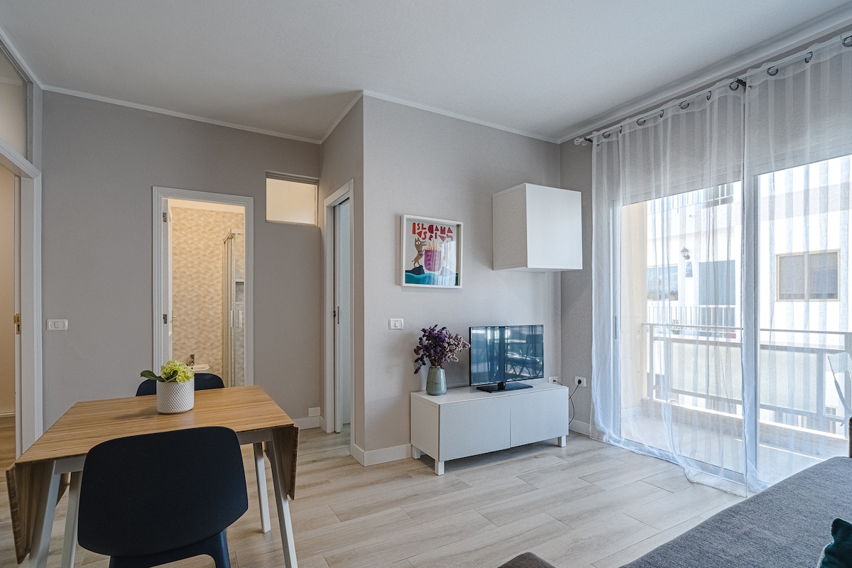 A light and airy living space features a wooden dining table paired with dark chairs. A modern white TV unit is positioned against a soft grey wall, with a television and tasteful decor displayed. Natural light filters through sheer curtains, illuminating the room.