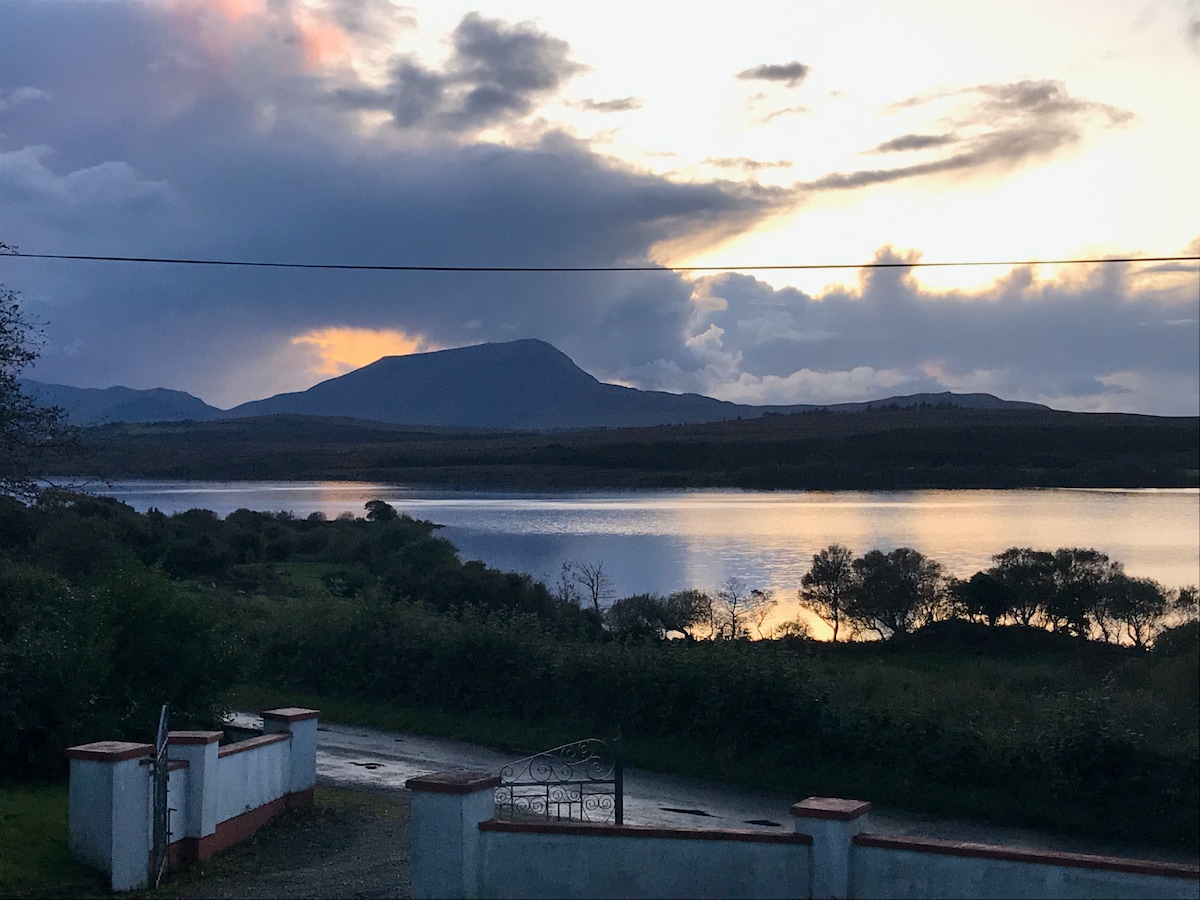 A serene landscape is captured, showcasing a lake reflecting the colors of the sunset. Rolling hills rise in the distance, under a cloudy sky. The entrance gate and driveway are visible in the foreground, leading towards the peaceful natural setting.