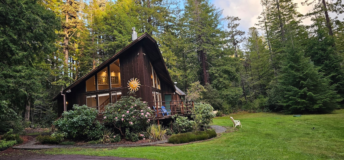 A two-story cabin is nestled among lush trees, featuring large windows that invite natural light. A welcoming garden with colorful flowers surrounds the entrance, while a deck extends from the front, complete with seating that overlooks the serene landscape.