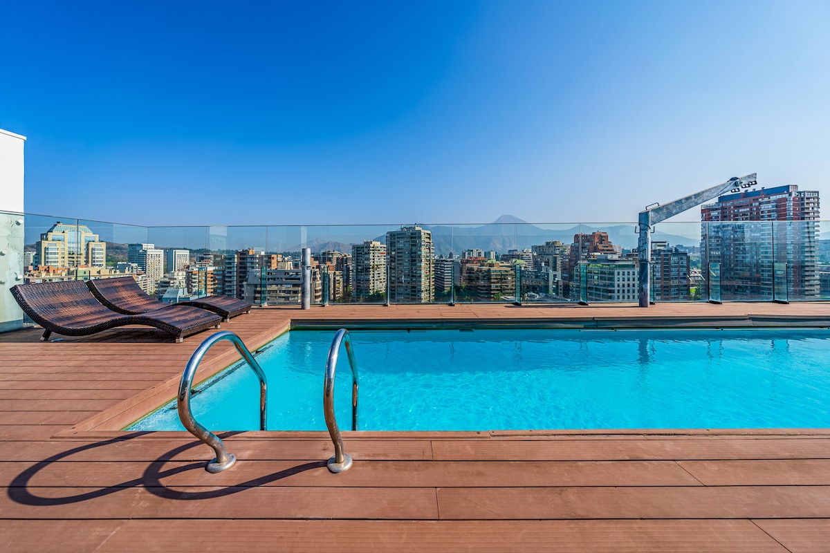 A rooftop pool provides a tranquil space with panoramic views of the city and mountains. The area features comfortable lounge chairs positioned beside the water, inviting relaxation under the clear blue sky.