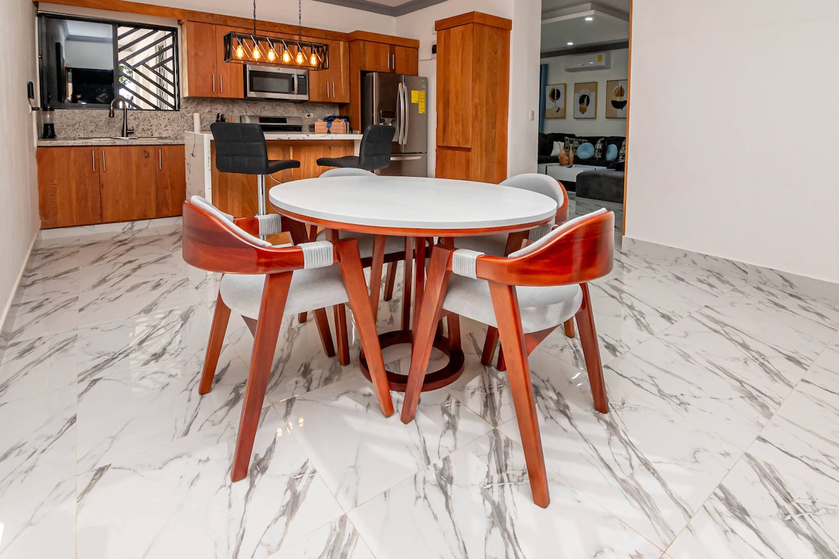 A modern dining area features a round table surrounded by four stylish chairs. Light wood accents complement the sleek marble floor, while the well-equipped kitchen is visible in the background, showcasing contemporary appliances and wooden cabinetry.