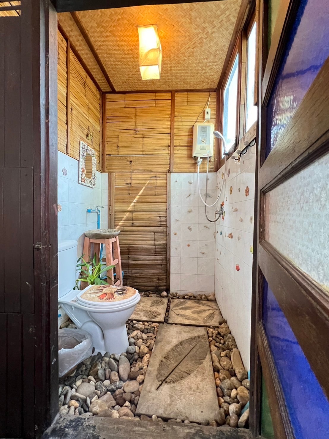 A private bathroom features a shower and a mix of natural stone and tiled surfaces. Large windows allow for natural light, while bamboo walls add a rustic touch. A small plant decorates the toilet area, enhancing the room's organic feel.