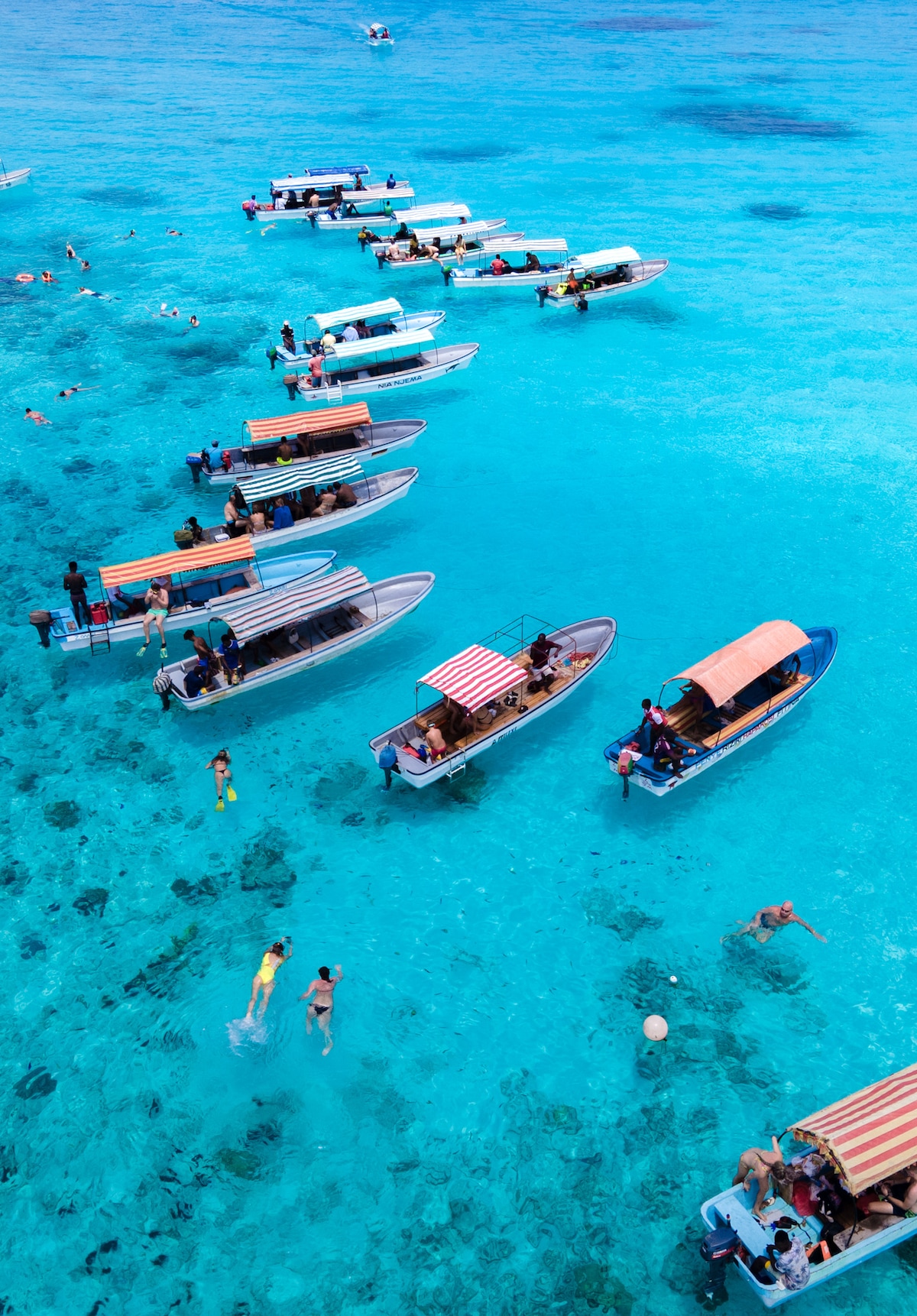Several boats are anchored in clear, turquoise waters, with people enjoying the sun and swimming nearby. The scene conveys a lively atmosphere with a mix of individuals engaged in water activities, highlighting the beauty of the surrounding environment.