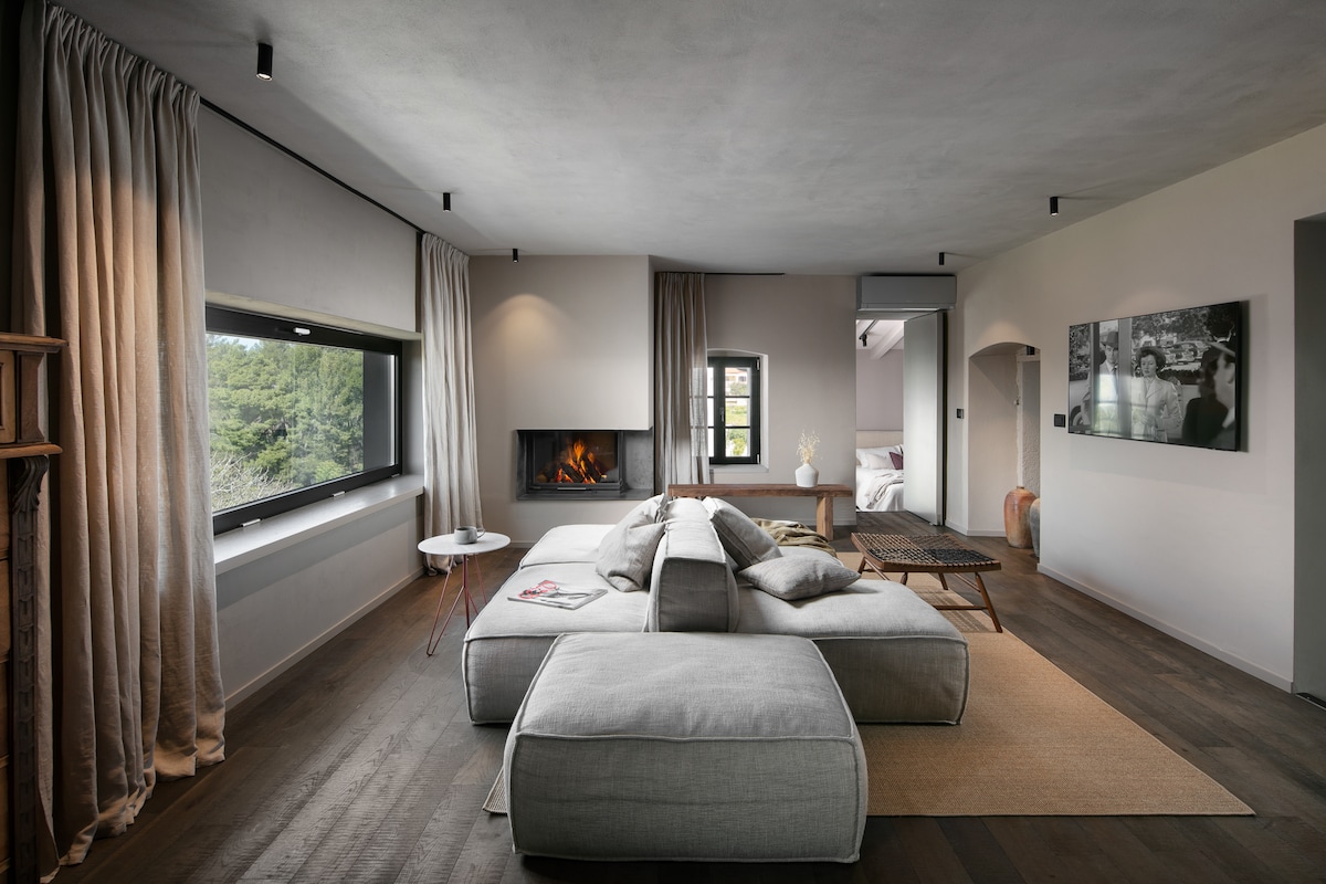 A spacious living area features a large sectional sofa, complemented by a sleek coffee table and floor mat. Natural light enters through a panoramic window, highlighting a contemporary fireplace. Soft curtains frame the window, enhancing a calm ambiance in the room.