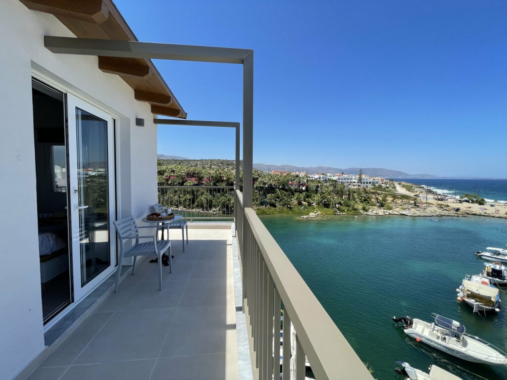 A balcony offers a scenic view of the coastal landscape, showcasing clear blue waters and a marina with boats. Two chairs and a small table are positioned on the balcony, emphasizing the inviting outdoor space. The surrounding area features lush greenery and a distant shoreline.