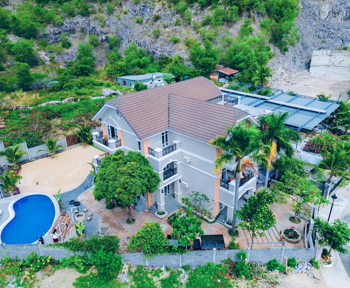 The villa is shown from an aerial perspective, highlighting its spacious layout. A private swimming pool is located beside the building, surrounded by lush greenery, with a sandy area and outdoor seating visible. The surrounding landscape includes rocky hills and well-maintained gardens.