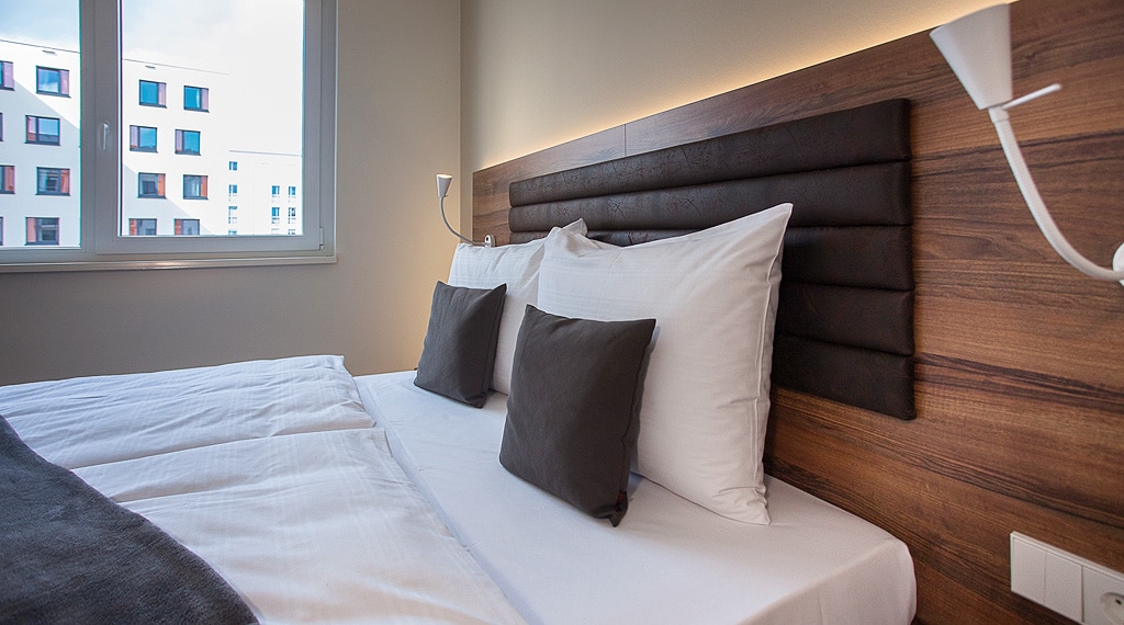 A modern sleeping area features a spacious bed with white linens and dark accent pillows. Warm wooden paneling runs along the headboard, while wall-mounted lamps provide soft illumination. A large window allows natural light to fill the room, framed by a simple, light-colored wall.