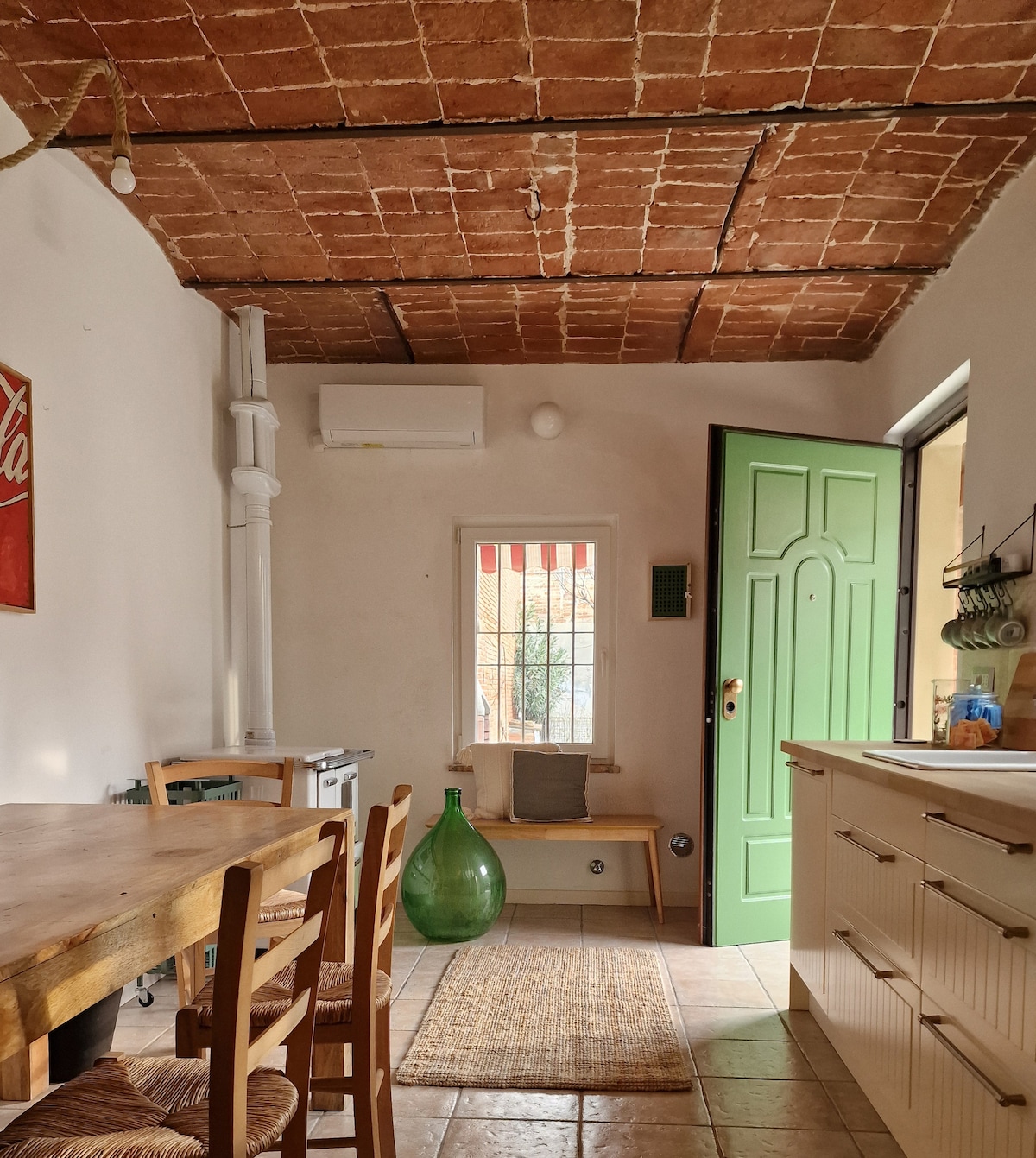 A bright kitchen area features a rustic wooden dining table accompanied by several wooden chairs. Natural light enters through a window with striped awnings. The green front door adds a pop of color, while a minimalist decor style is reflected in the clay-tiled ceiling.