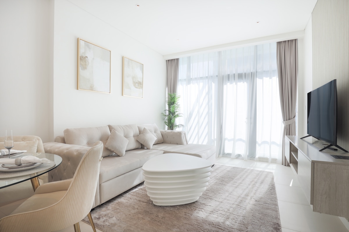 A modern living area is presented with a plush sectional sofa and a unique oval coffee table. Natural light fills the space through large floor-to-ceiling windows, while soft grey curtains frame the view. A dining table with elegant chairs is visible, enhancing the airy ambiance.