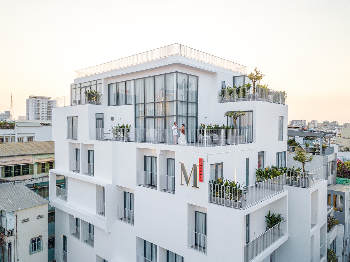The white multi-story building features large glass windows and a modern design. Lush greenery adorns the balconies, and the prominent 'M' signage is visible. A spacious rooftop terrace provides an inviting outdoor space, with city views in the background.