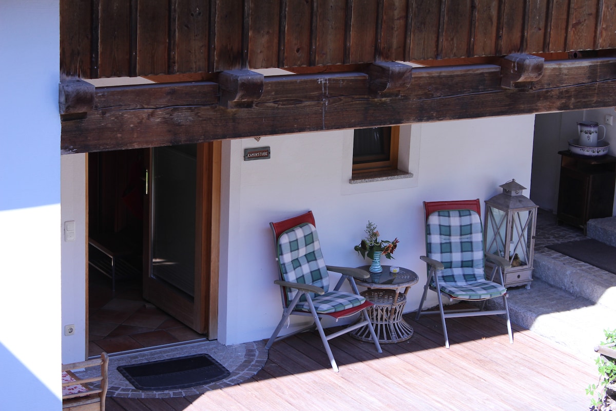 A cozy outdoor seating area features two plaid chairs facing a small round table. A lantern stands nearby, and a flower vase adds a touch of color. The entrance is framed by rustic wooden beams, creating a welcoming atmosphere.