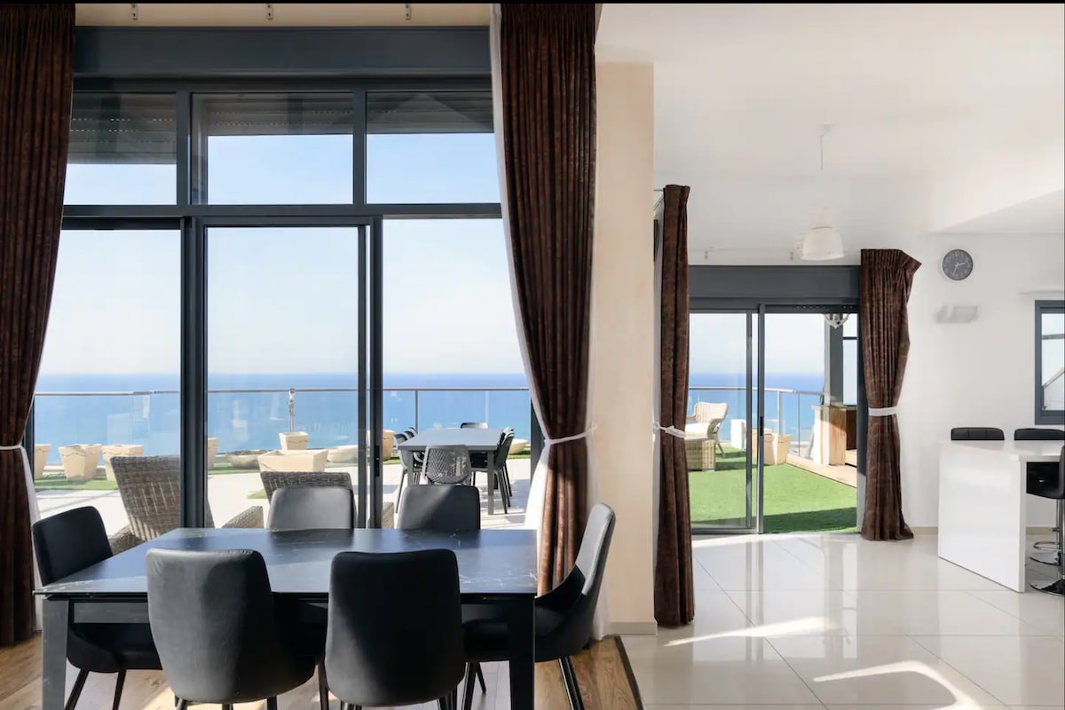 A spacious dining area is visible, featuring a dark table with black chairs. Large glass doors provide stunning ocean views and lead to an expansive outdoor terrace. Natural light fills the space, complemented by window coverings that frame the scenic vista.