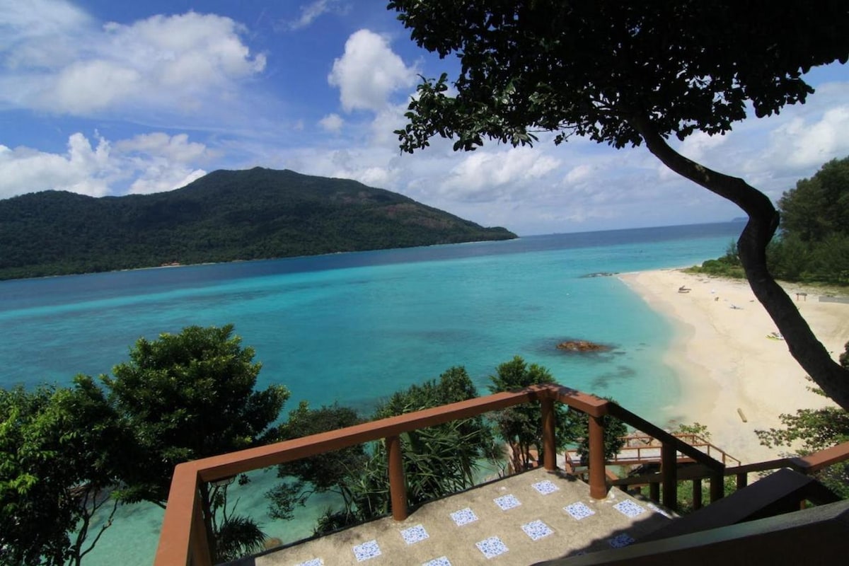 A scenic view features vibrant turquoise waters meeting the sandy beach, framed by lush greenery and gentle hills in the distance. The expansive sky showcases fluffy white clouds, enhancing the natural beauty of the coastline. A wooden balcony offers a vantage point to appreciate the surroundings.