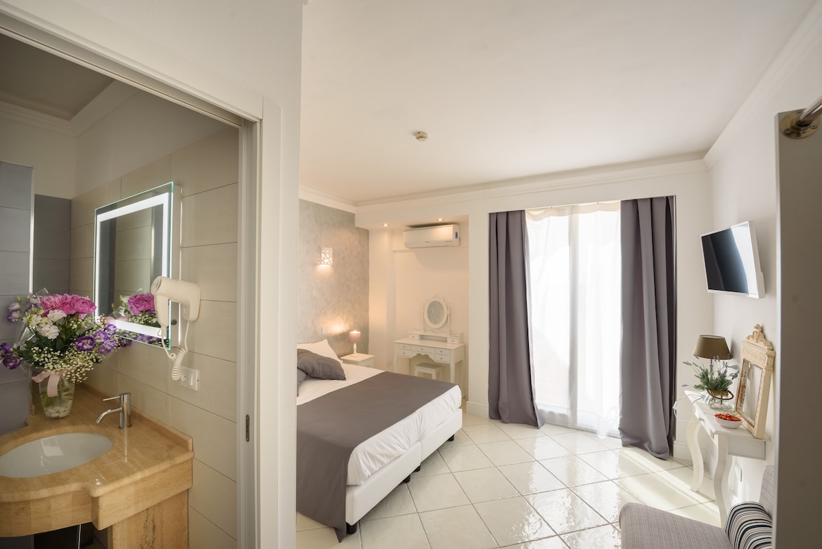 A spacious room features a neutral color palette with a comfortable bed centered in the layout. Natural light filters through large curtains, illuminating a cozy seating area and a small desk. A stylish bathroom with a vanity is visible, adorned with fresh flowers.
