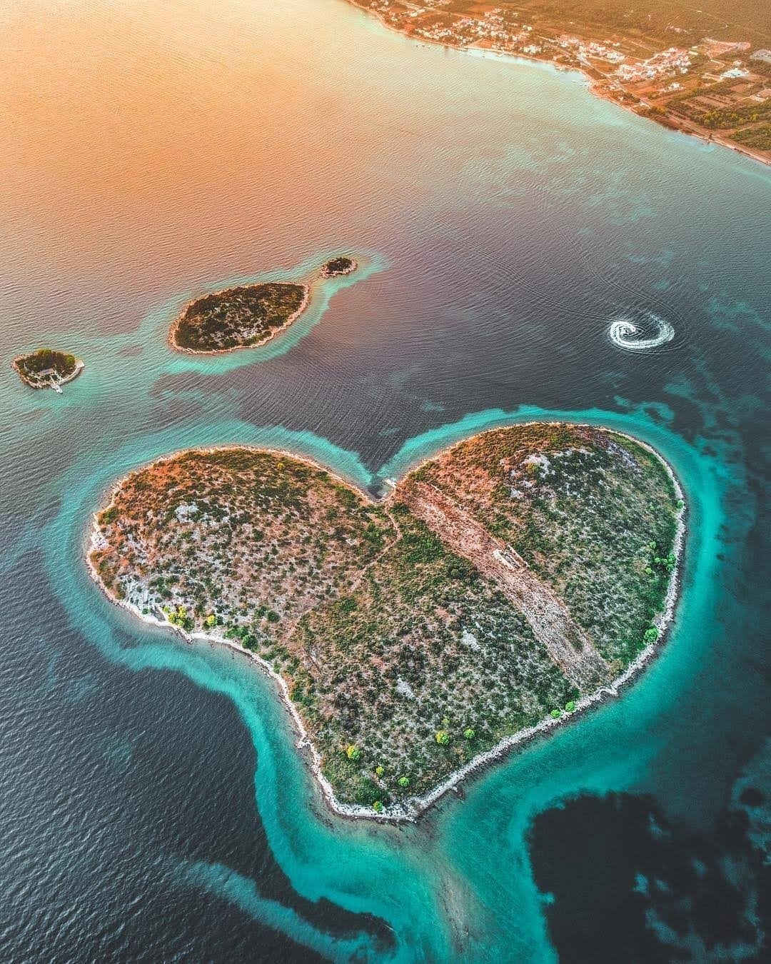 An aerial view captures a heart-shaped island surrounded by clear blue waters. Smaller islands are visible nearby, with gentle waves creating ripples around the heart-shaped landform. The soft hues of sunset enhance the natural beauty of the scene, blending the island's greenery with the water's vibrant colors.