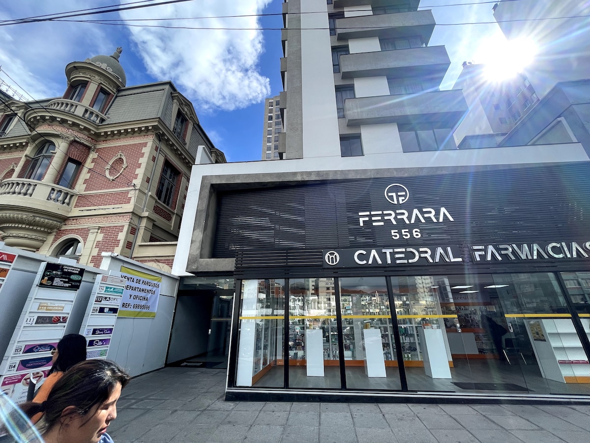 The image captures a vibrant urban scene featuring a modern building with a prominent storefront displaying the name 'FERARRA' and 'CATEDRAL FARMACIA'. In the background, a historic structure with intricate architectural details is observable. A clear blue sky enhances the overall atmosphere.
