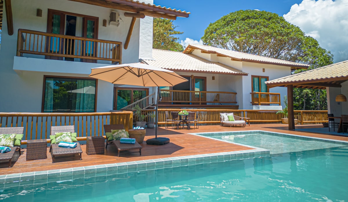 A spacious outdoor area features a tranquil infinity pool surrounded by sun loungers and an umbrella. The two-story house is set against a blue sky with clouds, showcasing wooden balconies and large windows that allow natural light to fill the space.
